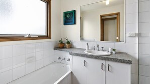 A modern bathroom with a bathtub, a sink, and a large mirror. There is a window with a wooden frame, and a painting is visible on the wall. - Ocean's Course (Lorne)