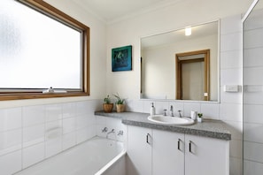 A modern bathroom with a bathtub, a sink, and a large mirror. There is a window with a wooden frame, and a painting is visible on the wall.