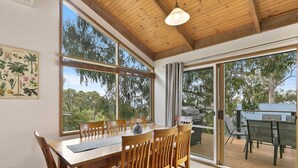 A dining area with a wooden table and chairs, large windows, and a view of the outdoors. - Ocean's Course (Lorne)