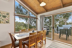 A dining area with a wooden table and chairs, large windows, and a view of the outdoors.