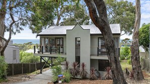A modern two-story house with a balcony, surrounded by trees and a view of the sea. - Ocean's Course (Lorne)