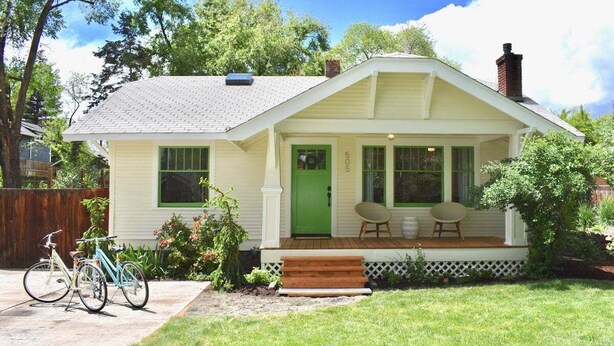 Une maison de plain-pied avec une porte verte, un porche doté de deux chaises et un vélo garé à l’extérieur.