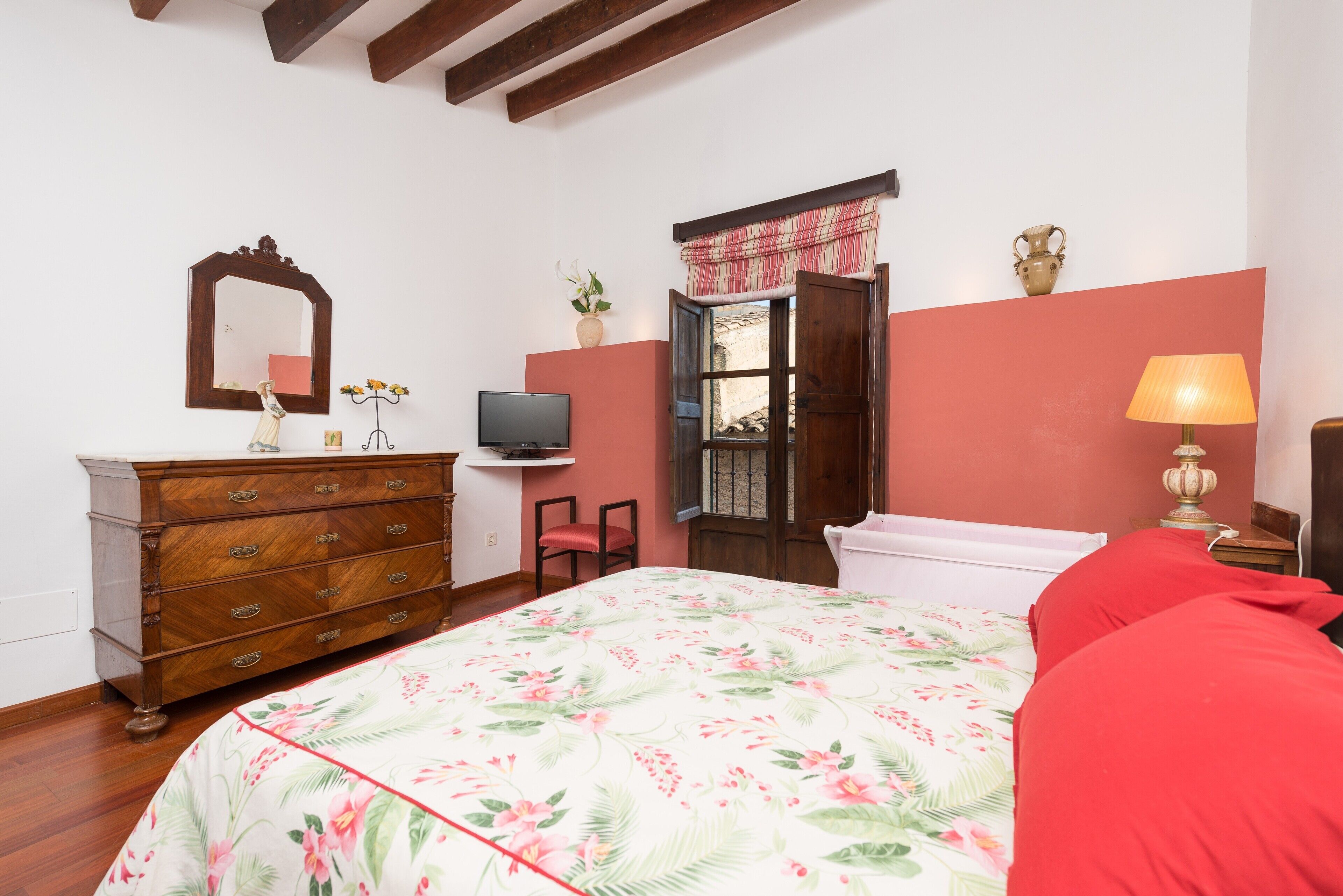 A bedroom with a wooden dresser, a television, a window with curtains, a bed with floral bedding, and a bedside lamp.