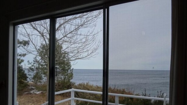 A room with large windows overlooking a beach. There are two white chairs on a wooden deck. - Oceanfront Carriage House Apt. w/ Spectacular Views (York)