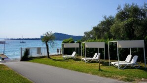 A lakeside area with three white sun loungers under white canopies, a paved walkway, and a view of the lake with boats and distant hills. - Villenpark Sanghen - Two-Room Apartment in Casa Centrale (Villaggio Sanghen)