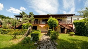 A two-story house with a stone pathway leading to the entrance, surrounded by lush greenery and a clear sky. - Villenpark Sanghen - Two-Room Apartment in Casa Centrale (Villaggio Sanghen)