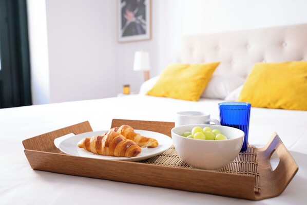 A breakfast tray with croissants, a bowl of grapes, and a blue glass on a bed with yellow pillows.