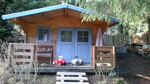 Eine Holzhütte mit blauem Dach, zwei Fenstern und einer Tür. Vor der Veranda stehen zwei Holzbanken und zwei Spielzeugautos.
