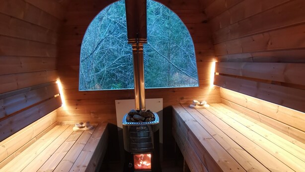 Ein Saunaraum mit Holzbänken, einer Zentralheizung und Blick auf Bäume durch ein halbrundes Fenster.