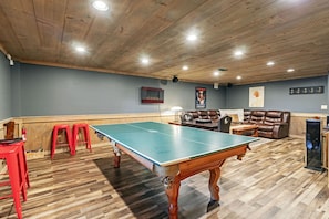 Una mesa de ping pong en una habitación con suelo de madera, un sofá de cuero marrón y un taburete rojo.