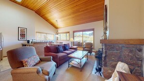 Un salon spacieux doté d’un haut plafond en bois, d’une cheminée et de grandes fenêtres offrant une vue sur l’extérieur.