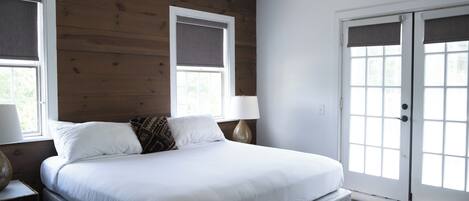 A bedroom with a large bed, a bench, two windows with blinds, and a door with glass panels.