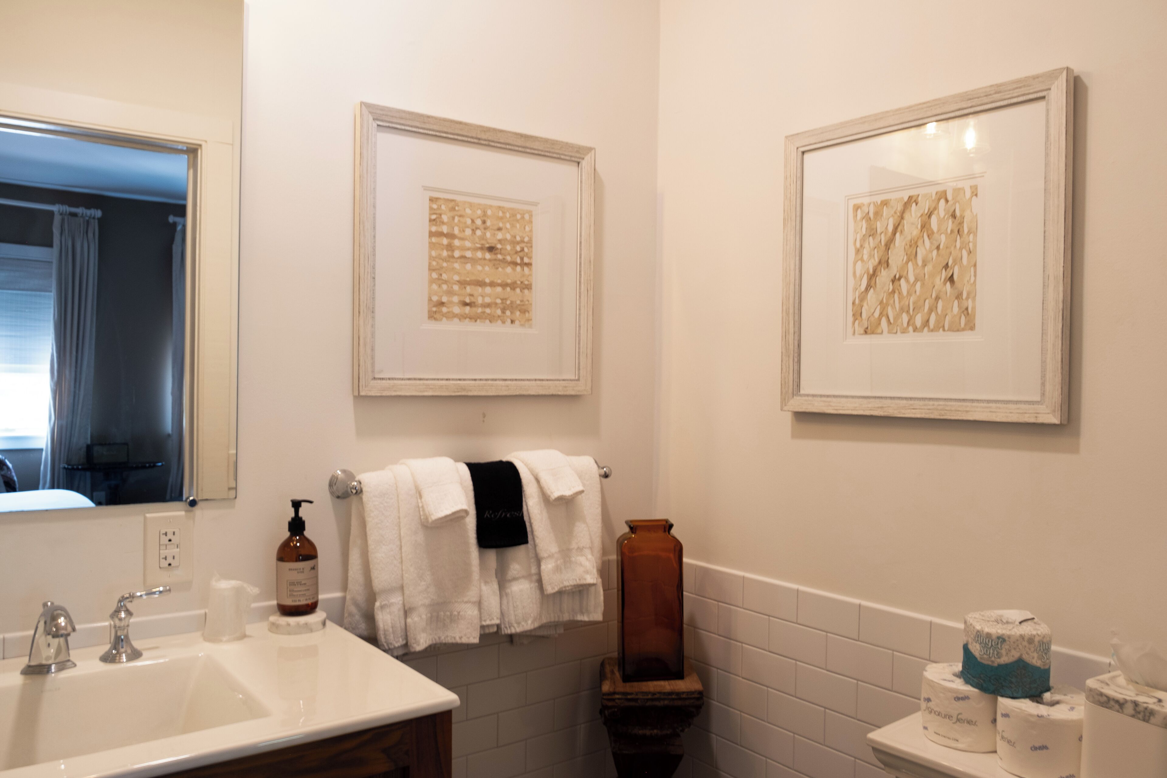 A bathroom with a sink, mirror, framed artwork, towels, and toilet paper.