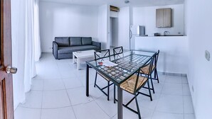 A modern living room with a grey sofa, a glass dining table with chairs, a white coffee table, a ceiling fan, and a kitchen area with a sink and cabinets.
