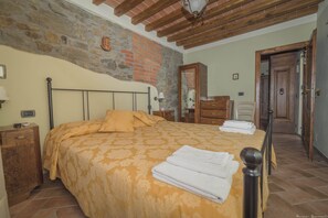 A bedroom with a bed, wooden headboard, a stone wall, a wooden dresser, a mirror, and a door.