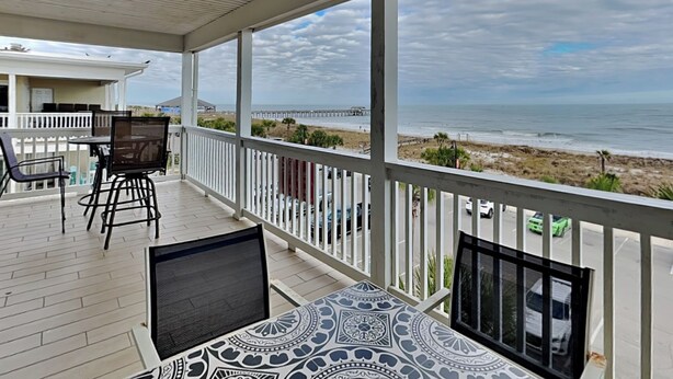 A covered outdoor area with a table and chairs, a view of the beach and ocean, and a pier in the distance. - Spacious w/ Fantastic Views, Large Balcony, Walkable Location! (Tybee Island)