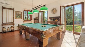 Sala de bilhar com mesa de sinuca, luminárias pendentes verdes e vista para o exterior através de grandes janelas.
