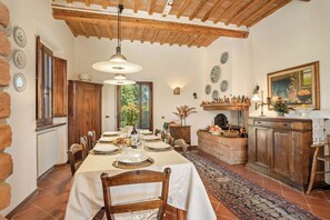 Une salle à manger avec une longue table dressée pour le repas, des poutres en bois au plafond et une cheminée agrémentée d’objets décoratifs.