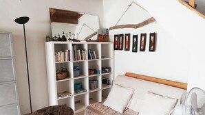 Een gezellige woonkamer met een houten tafel, een witte bank met kussens, een boekenplank vol boeken en decoratieve objecten, en een ventilator aan de muur.
