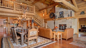 Interior espacioso de una cabaña de troncos, con comedor, chimenea y una escalera que sube a un nivel superior.