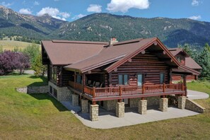 A log cabin with a stone foundation, a wooden deck, and a view of mountains. - Red Feather Lodge | Luxury Log Cabin for 28 (Big Sky)