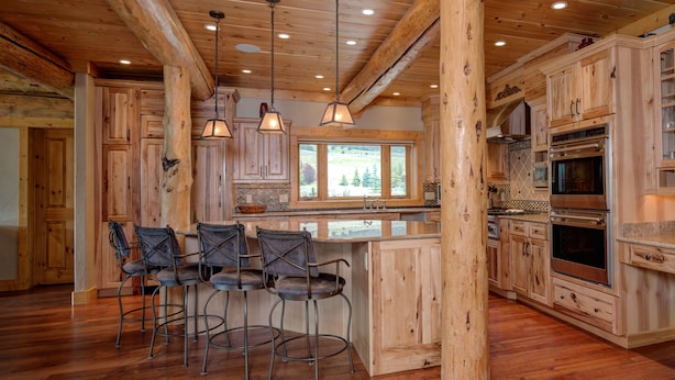 Interior de una cabaña de madera con cocina, que incluye armarios de madera, una zona de bar con taburetes y un ventanal con vistas a un paisaje pintoresco.
