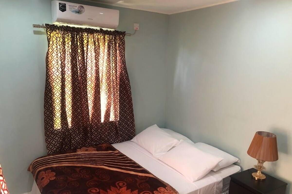 A single bed with a white mattress and pillows, a patterned bedspread, a bedside lamp, a dark nightstand, and an air conditioning unit mounted on the wall.