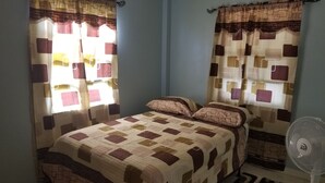 Un dormitorio con cama, cortinas, alfombra y ventilador de pie.