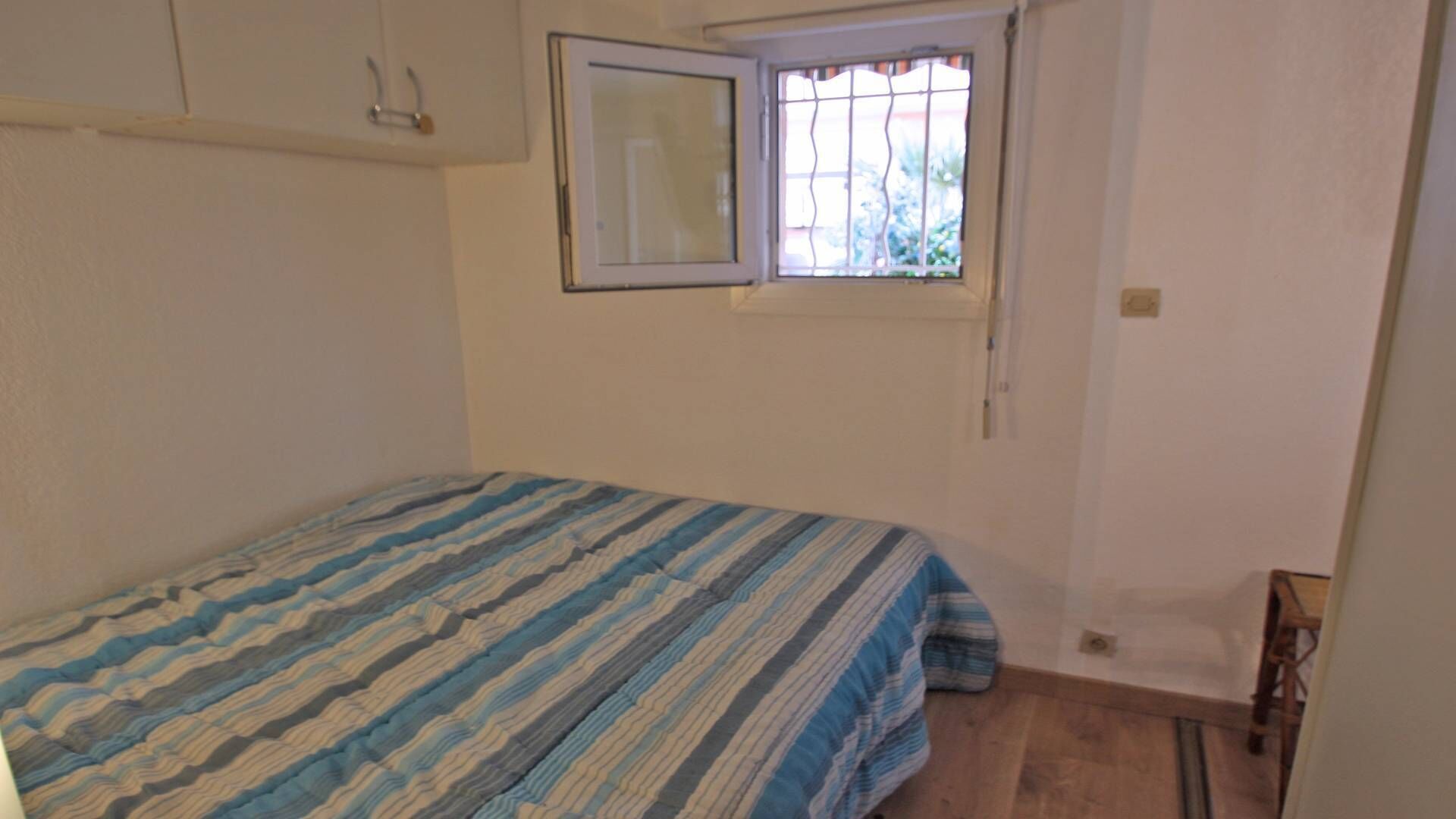 Una habitación pequeña y escasamente amoblada, con una cama individual, una ventana con rejas y una pared blanca y sin adornos.