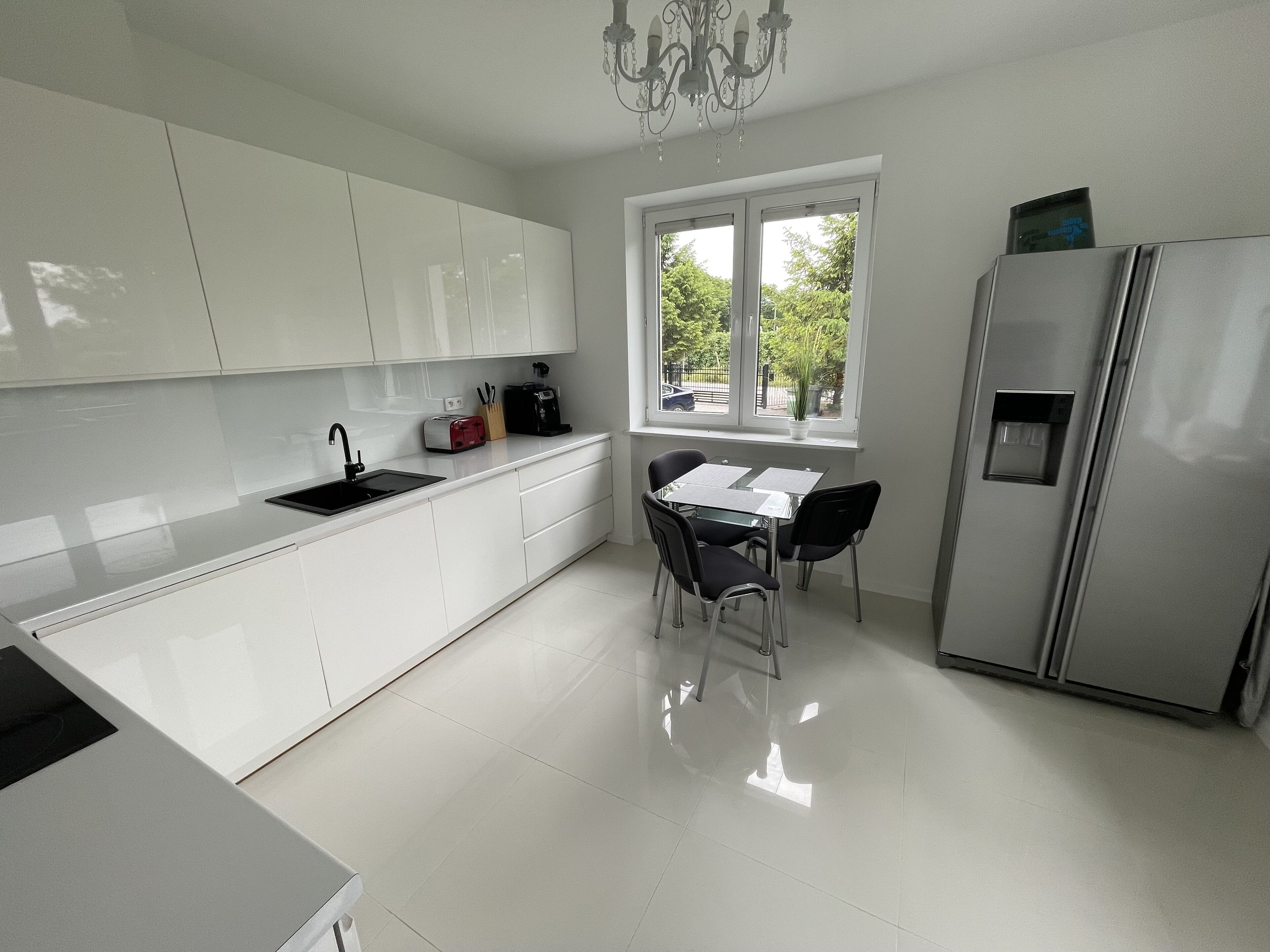 A modern kitchen with white cabinets, a stainless steel refrigerator, a dining table with chairs, and a window with a view of greenery outside.