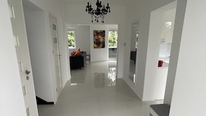Een moderne, goed verlichte hal met een kroonluchter, die leidt naar een zithoek met een bank en een kleurrijk schilderij.