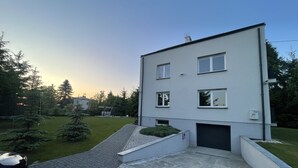 Een modern woonhuis met twee verdiepingen, een plat dak, een overdekte oprit en een kleine voortuin met gazon en enkele bomen.