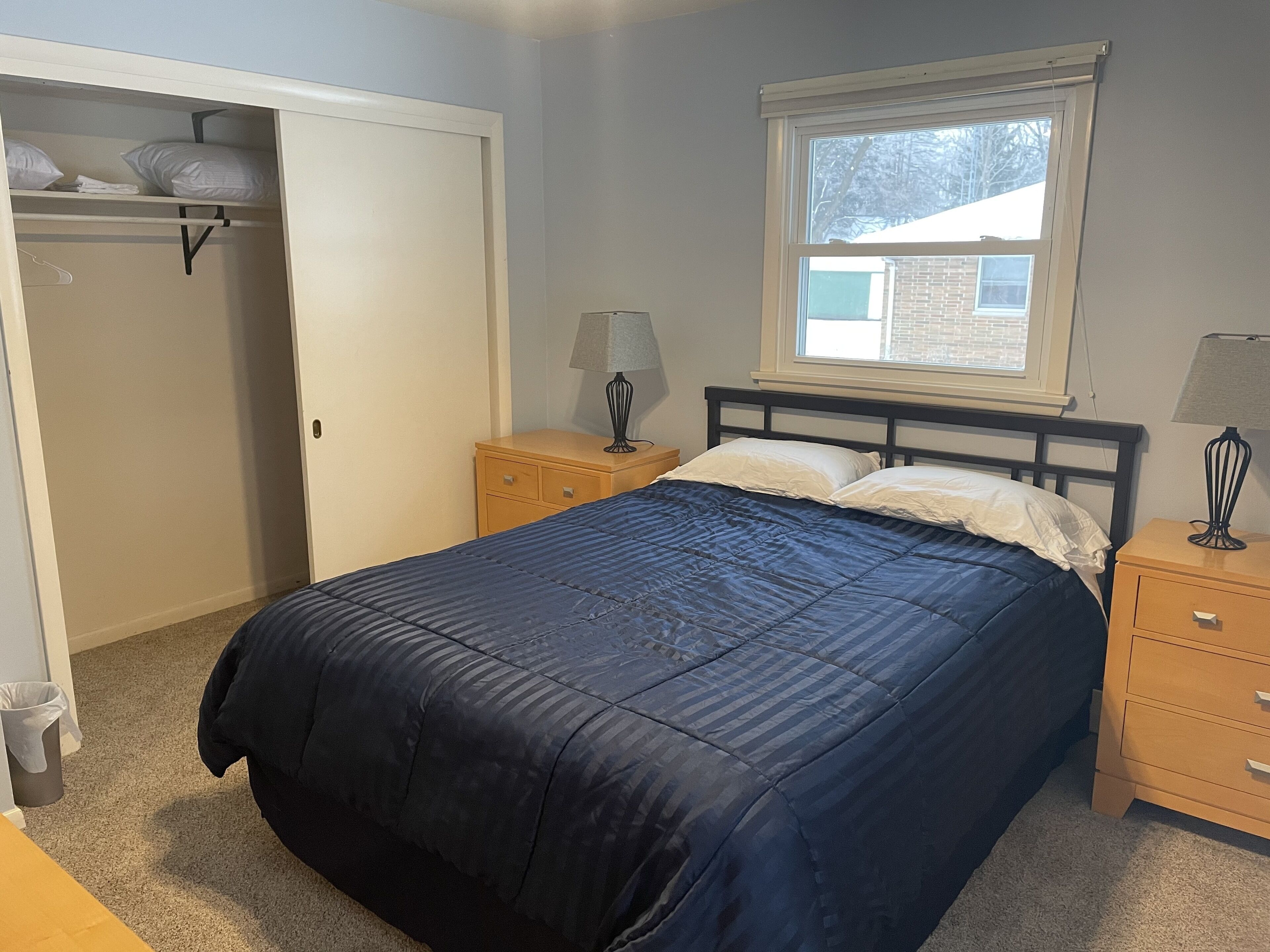 A bedroom with a bed, a nightstand, a lamp, a window, and a closet.