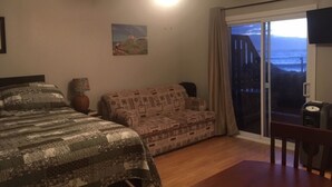 A bedroom with a bed, a sofa, a ceiling fan, a painting, and a view of the ocean through a sliding glass door. - Robbins by the Sea Rental - Robbins by the Sea 2 unit (Bonavista)