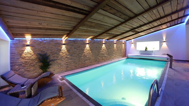 Une piscine intérieure avec un mur en pierre et un plafond en bois, équipée de chaises longues et d’une petite fenêtre.