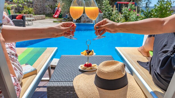 A poolside setting with two people toasting with orange juice glasses, a book, a hat, and a fruit plate on a table. - Apartments My Przno - Apartment with Side Sea View 4 (Przno, Budva)