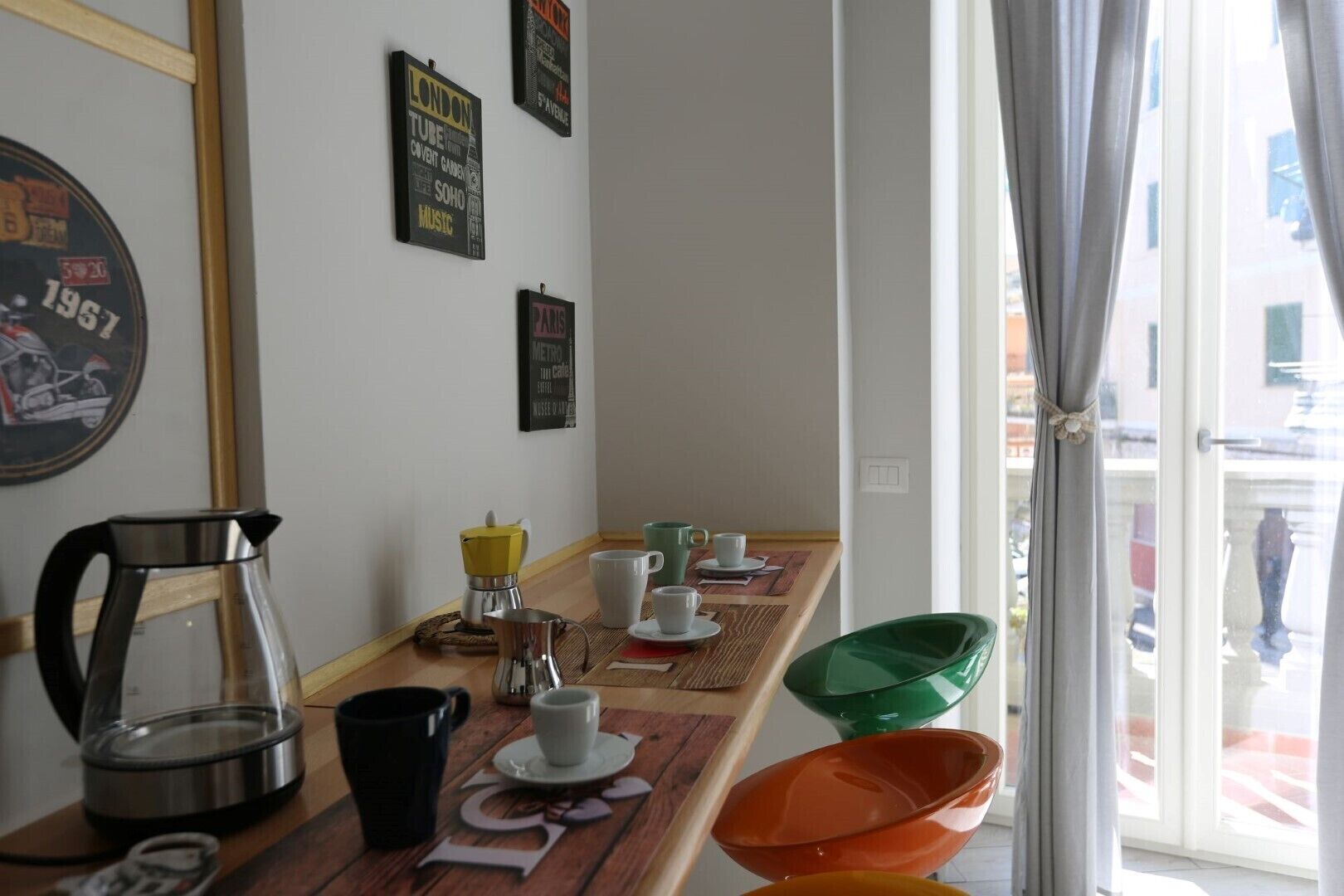 Une table de petit-déjeuner avec du café et du matériel pour préparer du thé, un mur orné d’affiches anciennes, et une vue sur la rue par la fenêtre.