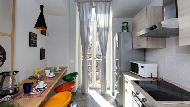 A compact kitchen with a microwave, stove, and a small refrigerator. There is a dining area with a wooden table and colorful chairs. - VILLA ROSA - Apartment n. 3 "VIOLA" (4 beds): (Napoli)