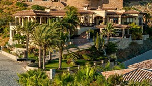 Une villa de style méditerranéen dotée d’une cour intérieure, de palmiers et d’un escalier en pierre menant à l’entrée.