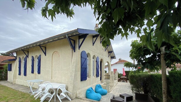 A building with a tiled roof, blue shutters, and a stone pathway leading to a patio area with outdoor seating and a small table. - Ref 006 - Charming Villa in the heart  of the resort (SOULAC SUR MER)