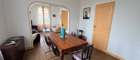 A dining area with a wooden table, chairs, and a blue vase. There is a sofa and a door in the background.