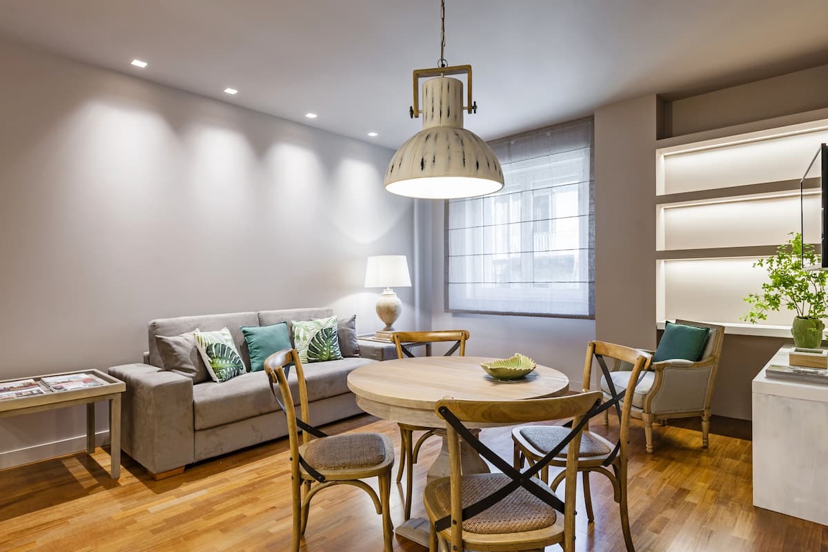 Una sala moderna con un sofá gris, una mesa de comedor de madera y sillas. Hay un ventanal con persianas, una lámpara colgante y una mesita de noche con lámpara.