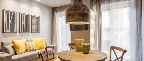 A modern living room with a round wooden table, two chairs, a sofa with yellow cushions, and a large hanging light fixture.