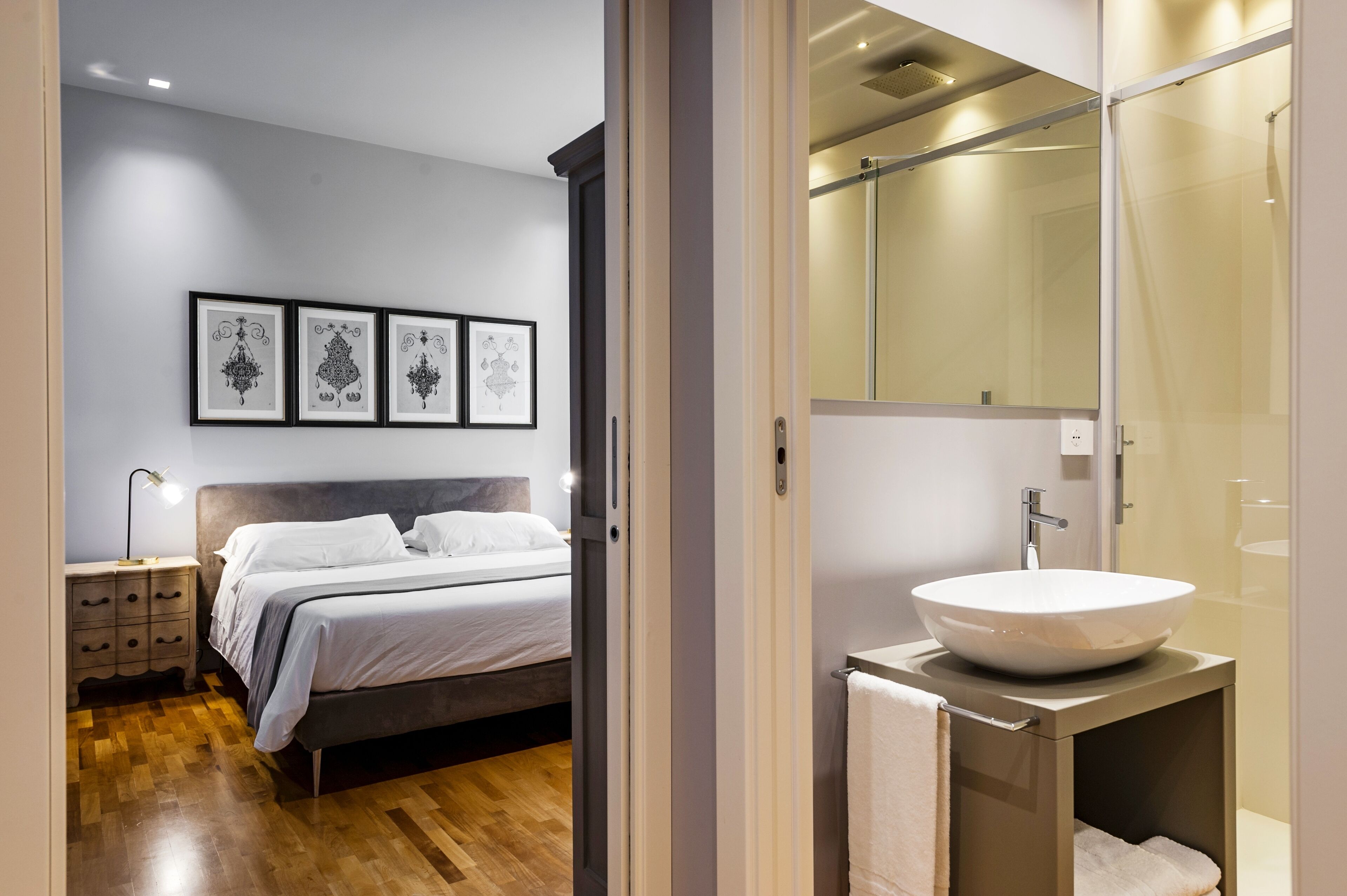 Un dormitorio con cama, mesita de noche y decoración en la pared. Junto a él, un baño con lavabo, espejo y ducha.
