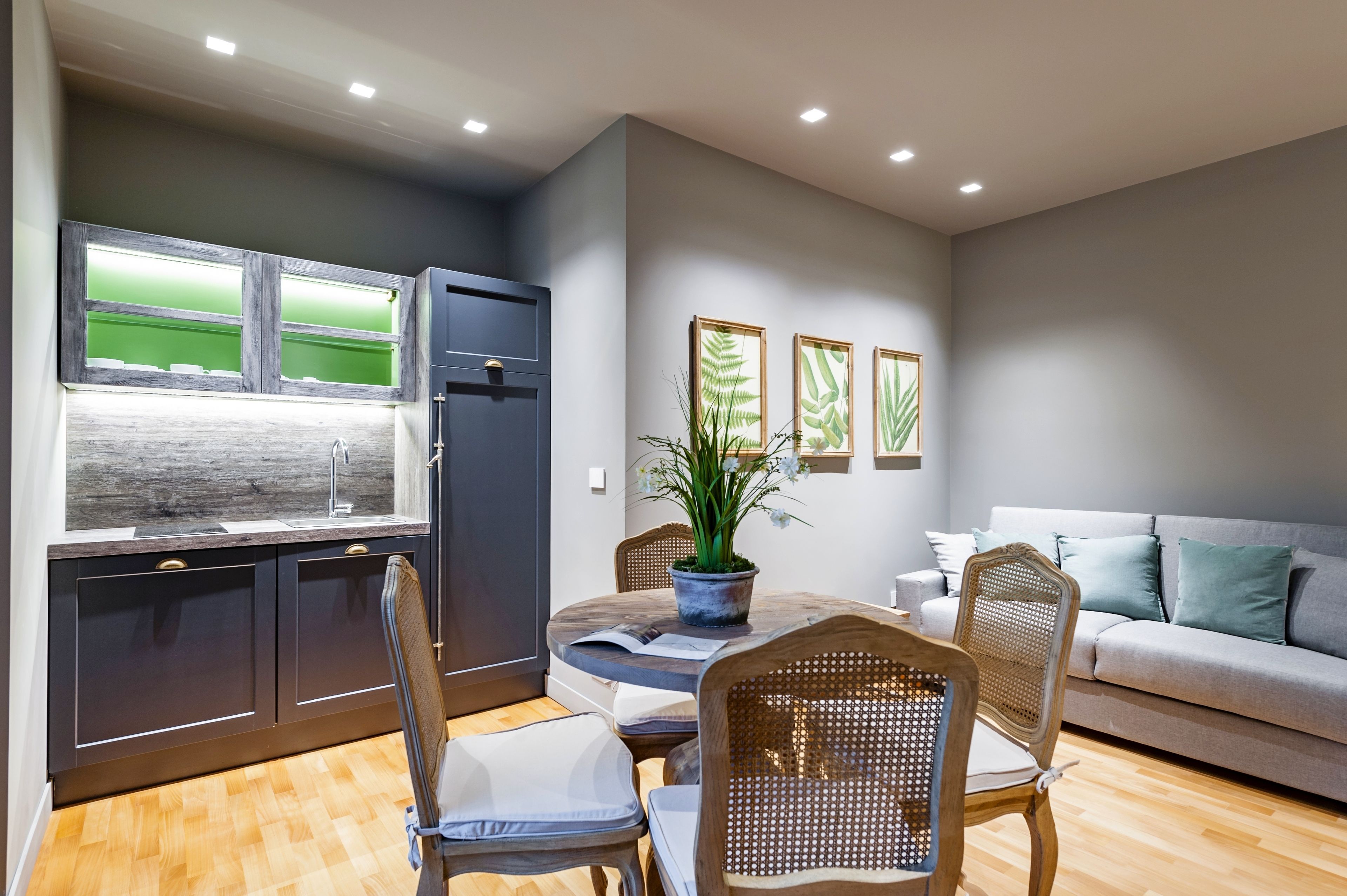 A modern kitchen with dark cabinets, a built-in refrigerator, and a window with a green view. There is a dining table with chairs and a potted plant. A grey sofa is in the living area with a framed plant on the wall.