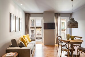 A modern living room with a sofa, dining table, and chairs. There is a TV mounted on the wall and a large window with curtains.