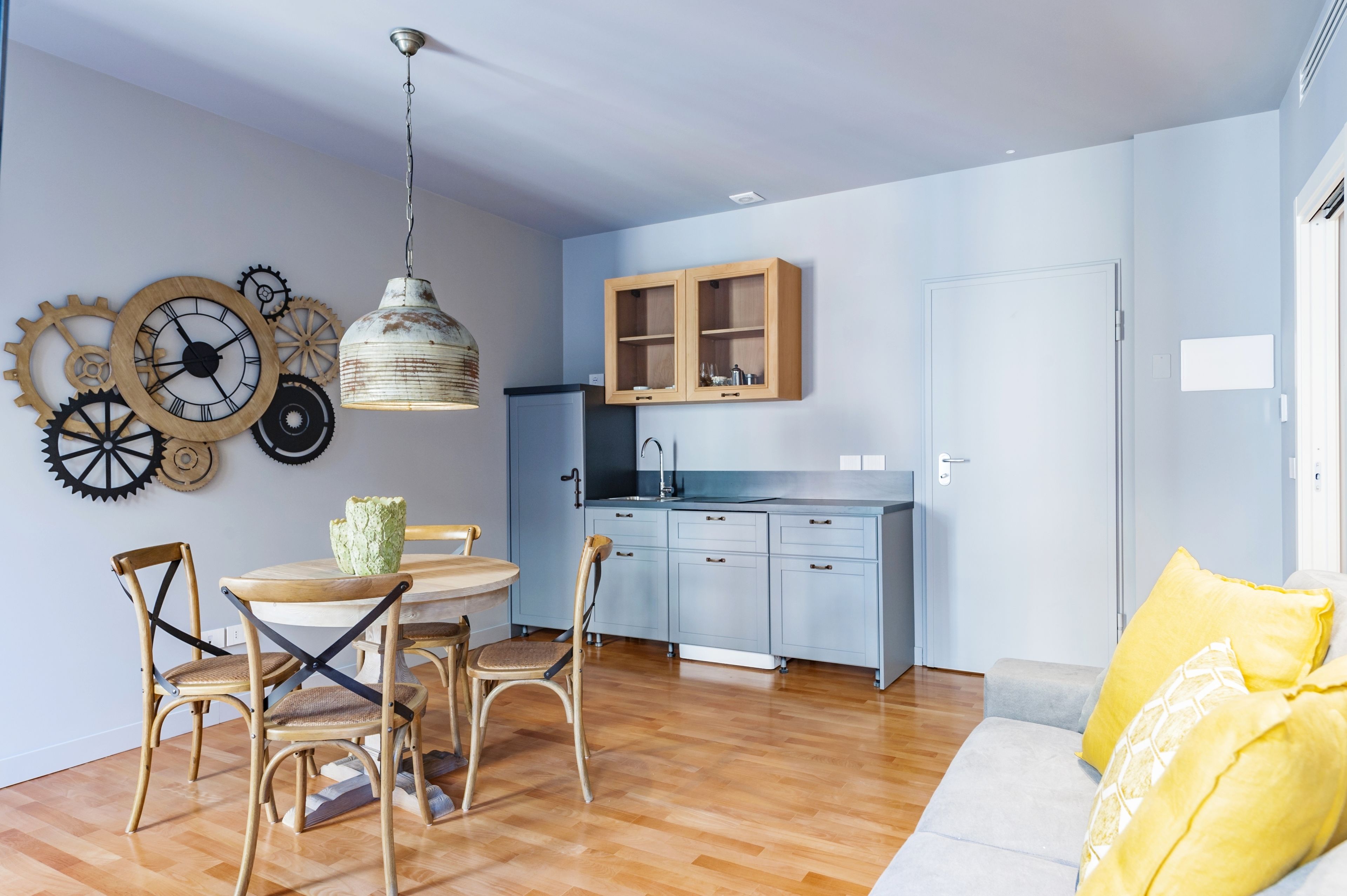 A modern kitchen with wooden cabinets, a dining area with a round table and chairs, and a living area with a sofa and cushions.
