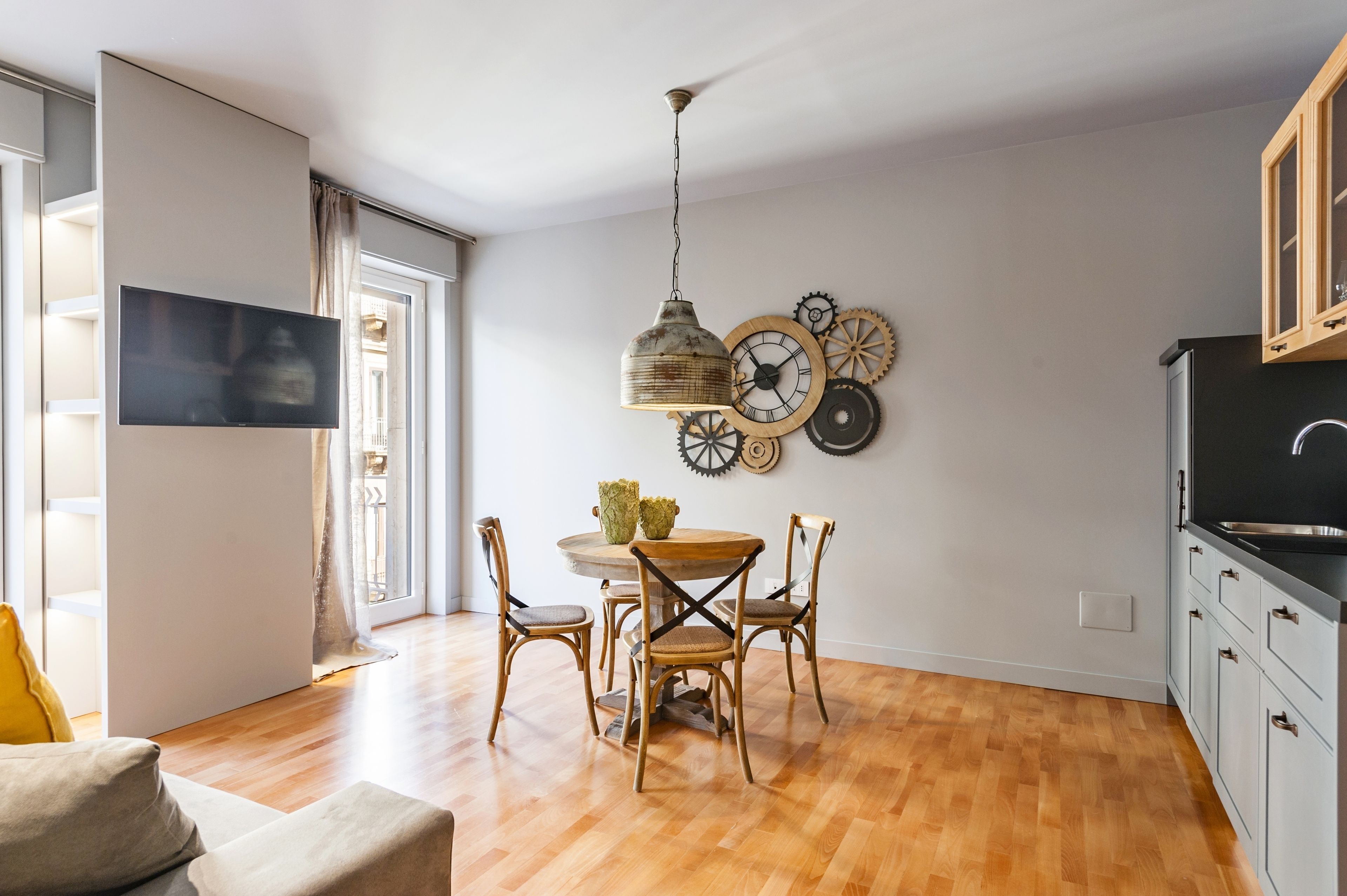 A modern kitchen with wooden cabinets, a dining area with a round table and chairs, and a wall-mounted TV.