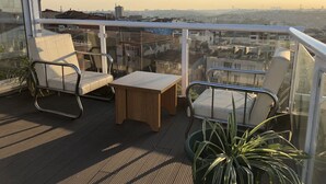 Un balcón con dos sillas y una pequeña mesa, con vista a un paisaje urbano con edificios y cielo despejado.