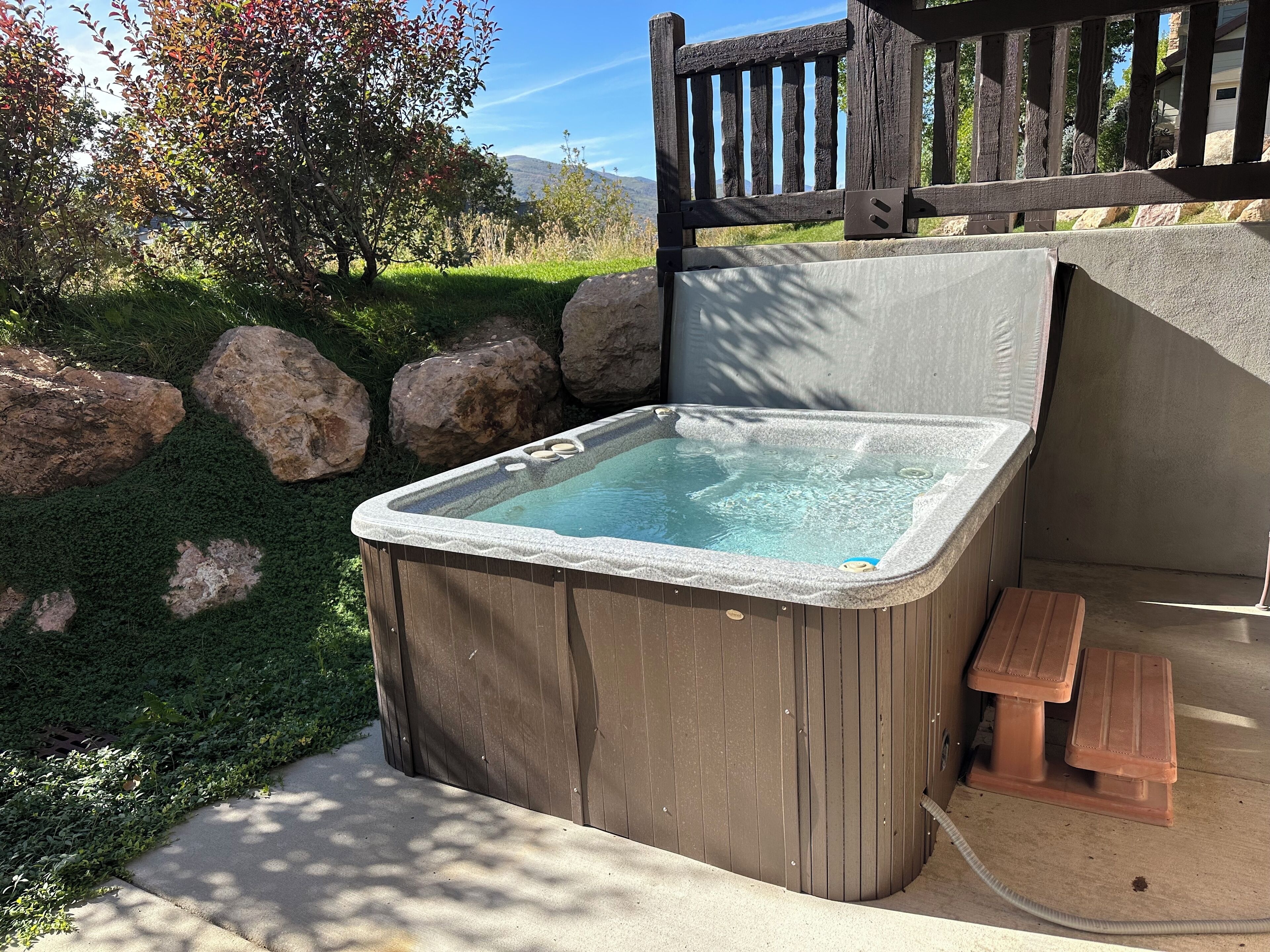 A hot tub with a wooden exterior, a small wooden bench, and a stone wall in the background.